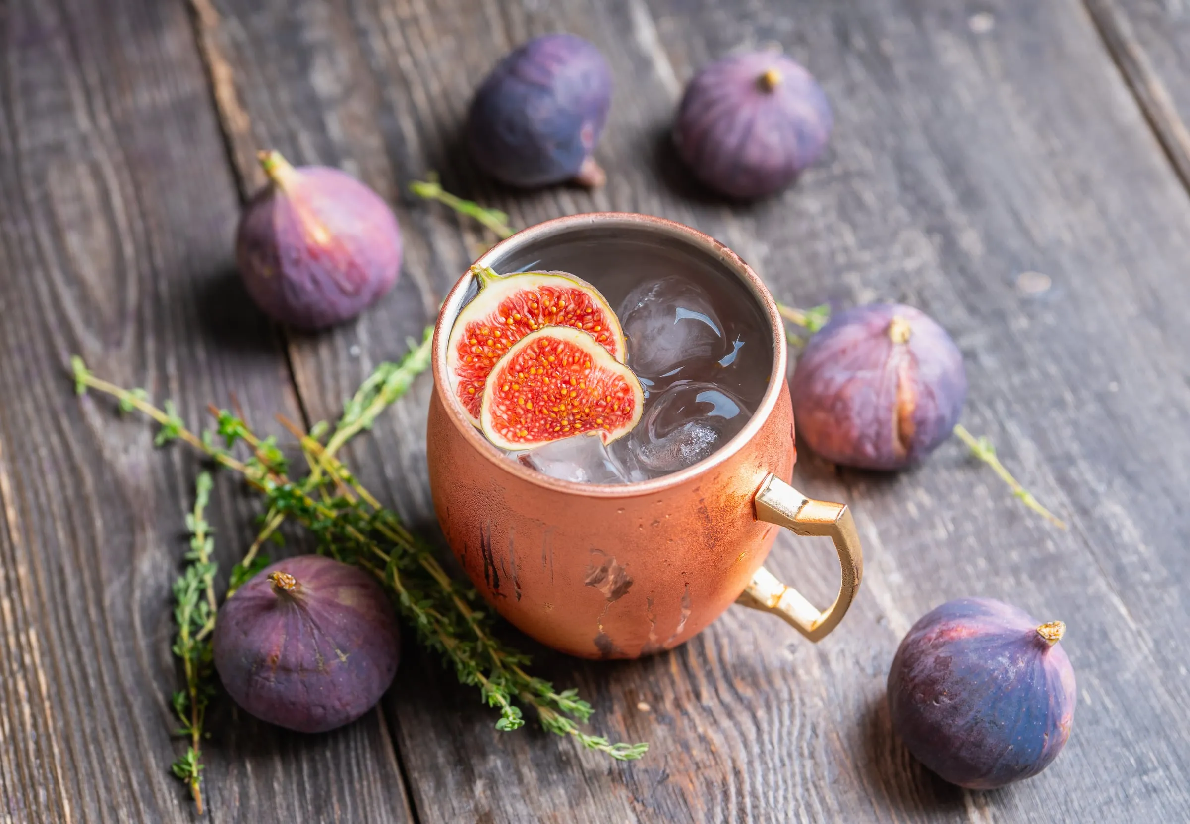 Fresh figs on an old wooden table around a copper mug with sliced fig, ice and thyme